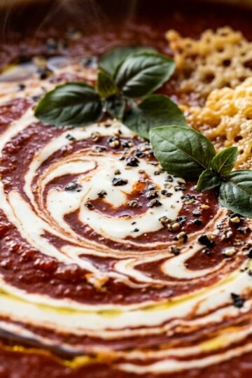 Ultra close-up macro texture of velvety creamy roasted tomato basil soup with cream swirl, fresh basil, and crispy parmesan crisps