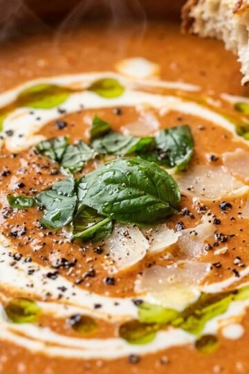 Ultra close-up macro of velvety roasted red pepper and tomato bisque in a bowl with cream swirl, fresh basil ribbons, and golden garlic croutons